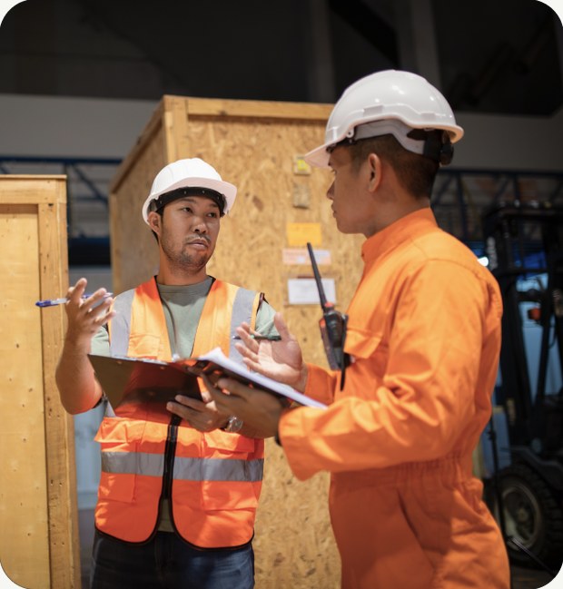 Two warehouse workers in hard hats reviewing shipment paperwork beside wooden crates