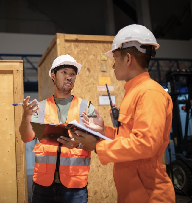 Two warehouse workers in hard hats and safety vests reviewing shipment paperwork beside wooden crates