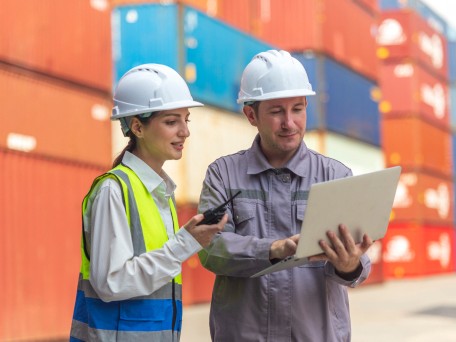 Two workers reviewing shipping documents on a laptop beside cargo containers